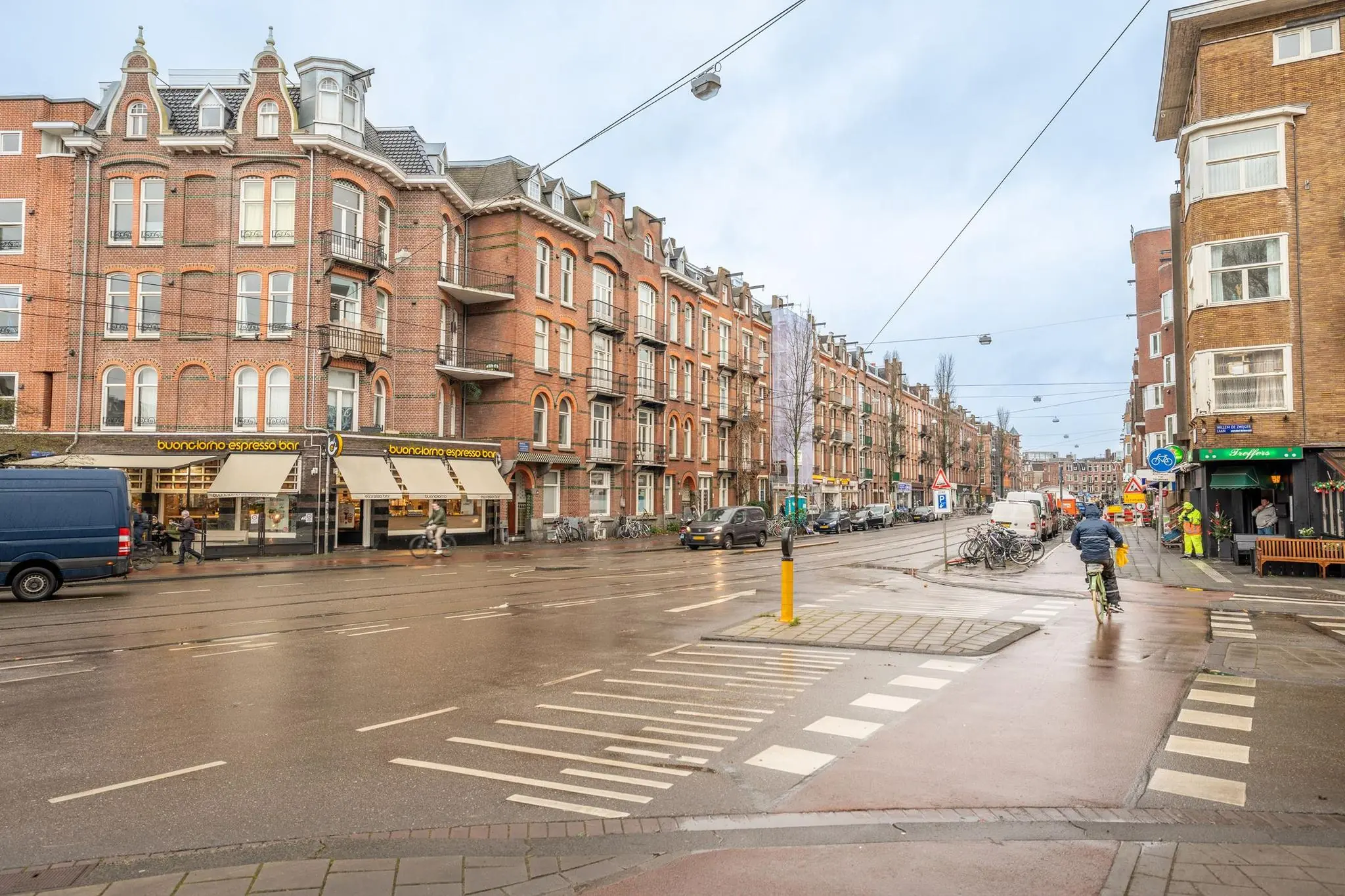 Straatbeeld van de Admiraal de Ruijterweg in Amsterdam met karakteristieke bakstenen panden, winkels en fietsers.